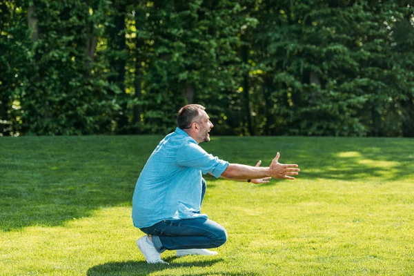 Seitenansicht eines glücklichen Mannes mit offenen Armen, der im Park hockt und wegschaut — Stockfoto