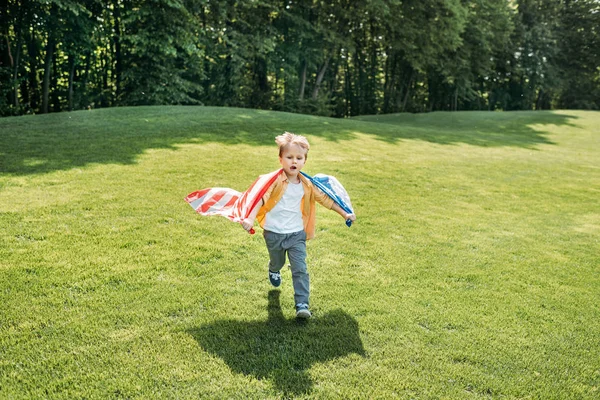 Entzückender kleiner Junge mit amerikanischer Flagge läuft auf Gras im Park — Stockfoto