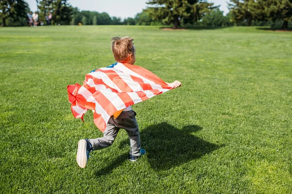 Vue arrière du petit garçon avec drapeau américain courant sur l'herbe dans le parc — Photo de stock