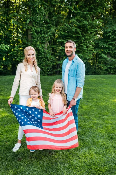 Familia feliz con dos niños sosteniendo la bandera americana y sonriendo a la cámara en el parque - foto de stock