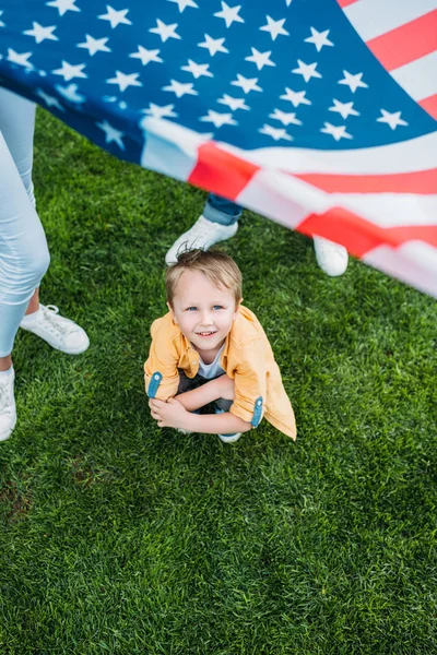 Recortado disparo de padres sosteniendo bandera americana y poco hijo agachándose en la hierba - foto de stock