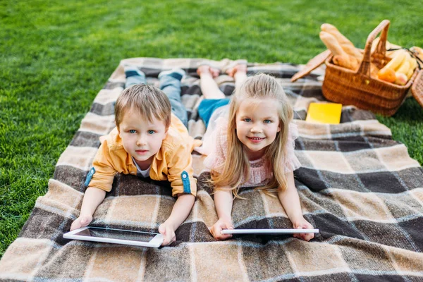 Mignons petits enfants à l'aide de tablettes numériques et souriant à la caméra tout en étant couché au pique-nique — Photo de stock