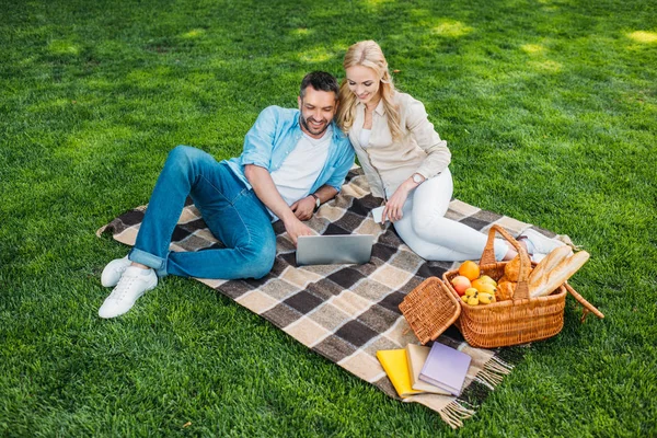 Vista de ángulo alto de la pareja feliz usando el ordenador portátil en el picnic a cuadros en el parque - foto de stock