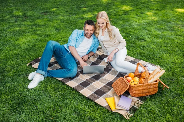 Vue grand angle de couple heureux à l'aide d'un ordinateur portable sur plaid au pique-nique dans le parc — Photo de stock