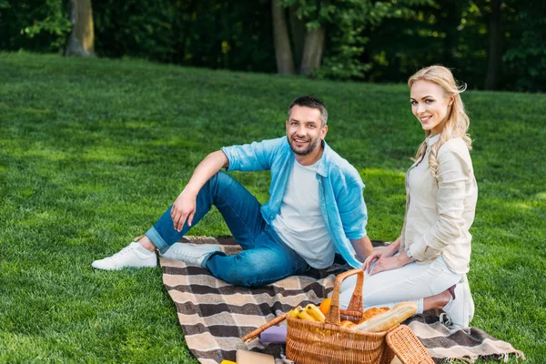 Feliz joven pareja sentado en cuadros y sonriendo a la cámara en el picnic en el parque - foto de stock