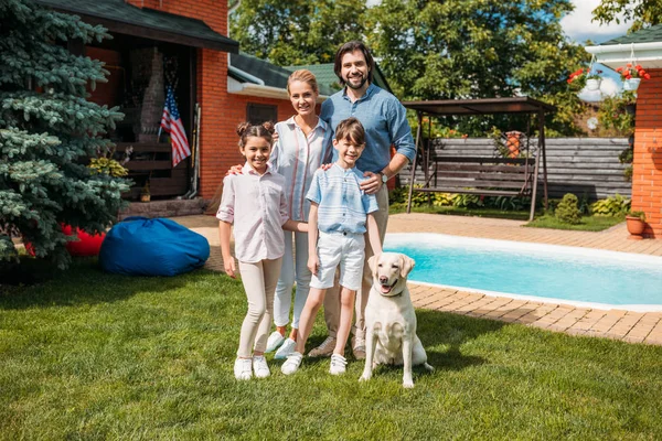 Весела сім'я з собакою-лабрадором, яка дивиться на камеру, стоячи на подвір'ї заміського будинку в літній день — стокове фото