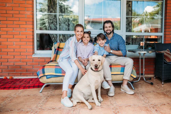 Sonriente familia labrador perro sentado en sofá juntos en casa de campo porche - foto de stock