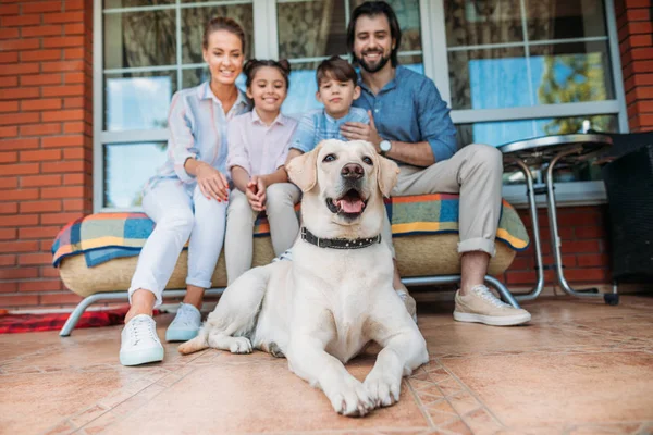 Улыбающаяся семья с собакой-лабрадором, сидящей вместе на диване на крыльце загородного дома — стоковое фото