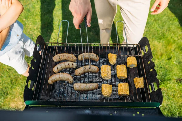 Cropped shot of son looking at father cooking sausages and corn on grill on backyard — Stock Photo