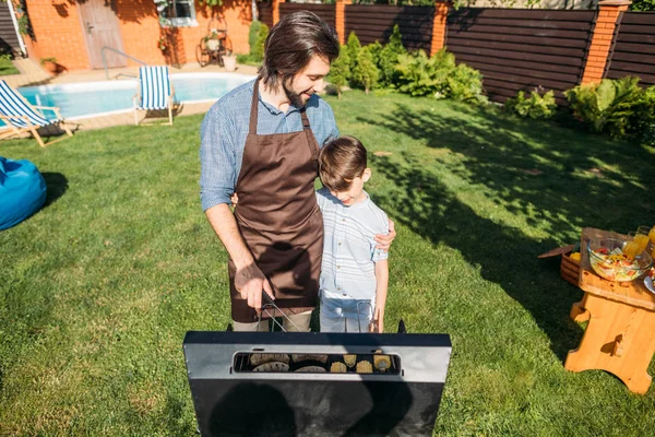 Син дивиться на батька, який готує ковбаски та кукурудзу на грилі на задньому дворі — стокове фото