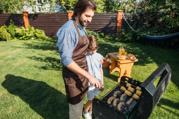 Hijo mirando padre cocinar salchichas y maíz en la parrilla en el patio trasero - foto de stock
