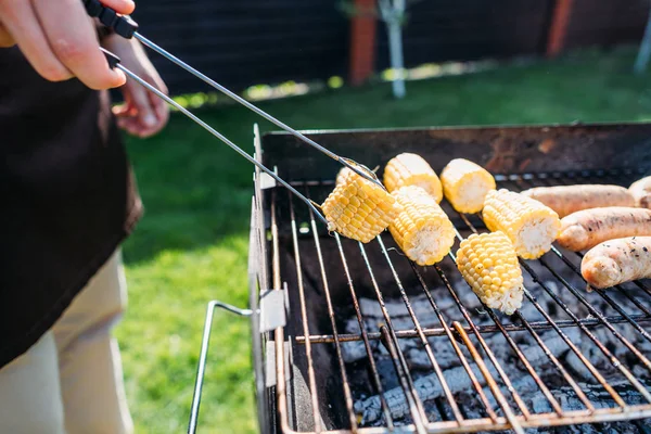 Partial view of man cooking corn and sausages on barbecue on summer day — Stock Photo