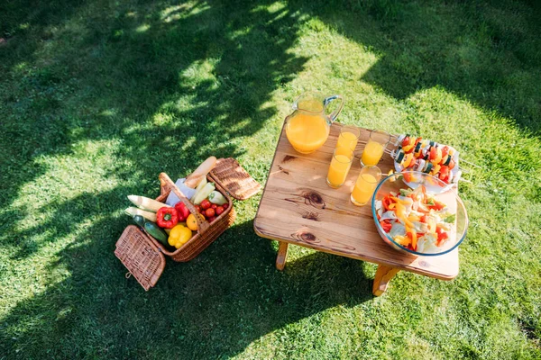 Vista superior de la cesta con comida para picnic, vasos de jugo y ensalada fresca en silla de madera en césped verde - foto de stock