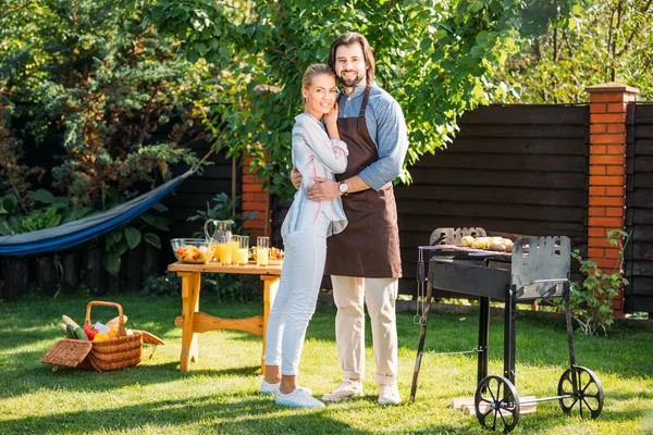 Sonriente pareja mirando la cámara teniendo barbacoa en el patio trasero en el día de verano - foto de stock