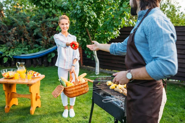 Vista parcial de la mujer sonriente con cesta de picnic dando pimiento al marido durante la barbacoa en el patio trasero - foto de stock