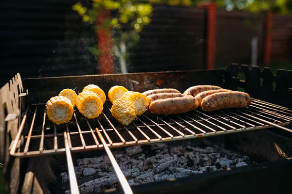 Close up view of cooking process of corn and sausages on grill on summer day — Stock Photo
