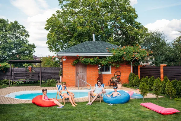 Familia descansando en sillas de playa y sillas de frijol cerca de la piscina en el patio trasero del campo en el día de verano - foto de stock