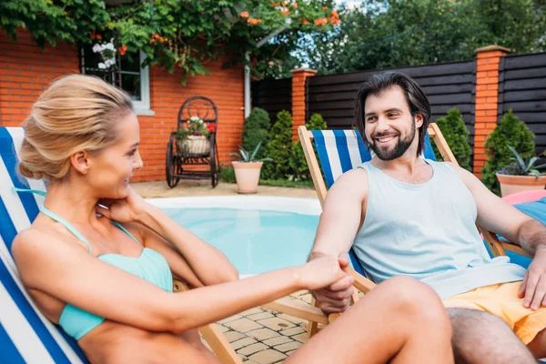 Пара на пляжних стільцях тримає руки, проводячи час біля басейну на задньому дворі в літній день — стокове фото