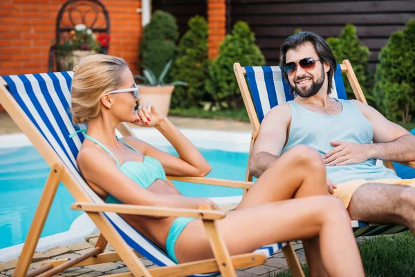 Sonriente pareja en sillas de playa pasar tiempo cerca de la piscina en el patio trasero en el día de verano - foto de stock