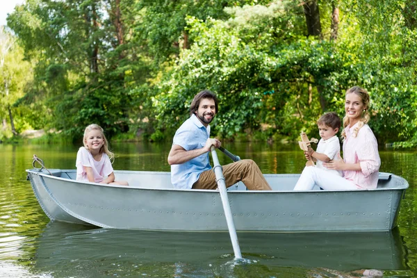 Vista lateral de sorrir jovem família equitação barco no rio no parque e olhando para a câmera — Fotografia de Stock