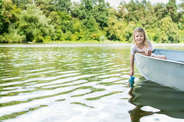 Linda criança lançando barco de origami de papel enquanto flutuava em barco no lago — Fotografia de Stock