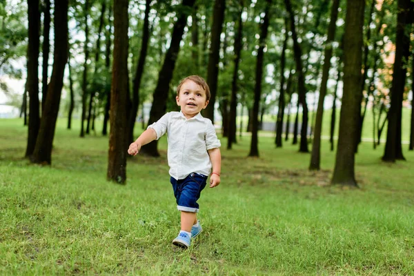 Petit enfant souriant en chemise blanche et short en denim couru par le parc — Photo de stock