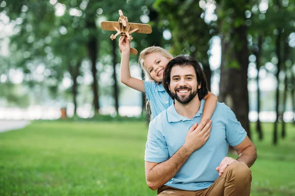 Adorable smiling daughter embracing happy father from behind and holding toy airplane at park — Stock Photo