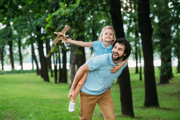 Sonriente hija piggybacking en feliz padre y jugando con juguete avión en parque - foto de stock