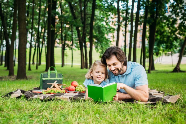 Handsome happy father and daughter lying on plaid in park and reading book at park — Stock Photo