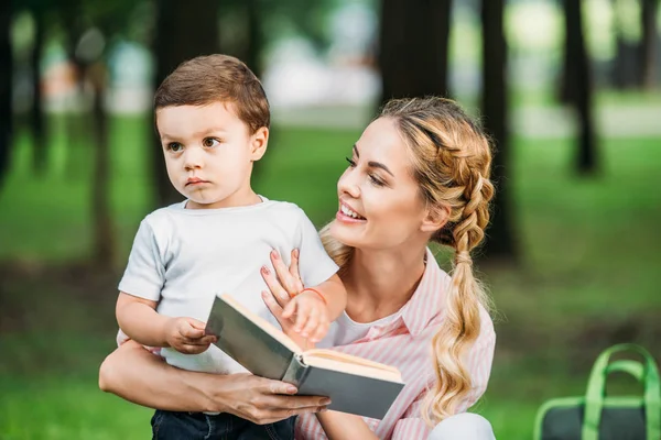 Mãe feliz com o pequeno filho triste leitura livro no parque — Fotografia de Stock