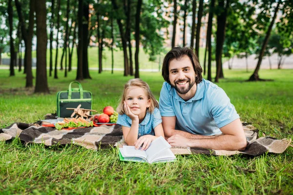 Gut aussehender glücklicher Vater und Tochter liegen auf Plaid im Park und lesen Buch im Park — Stockfoto