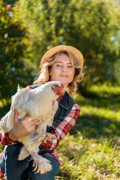 Smiling woman in straw hat holding white chicken outdoors — Stock Photo