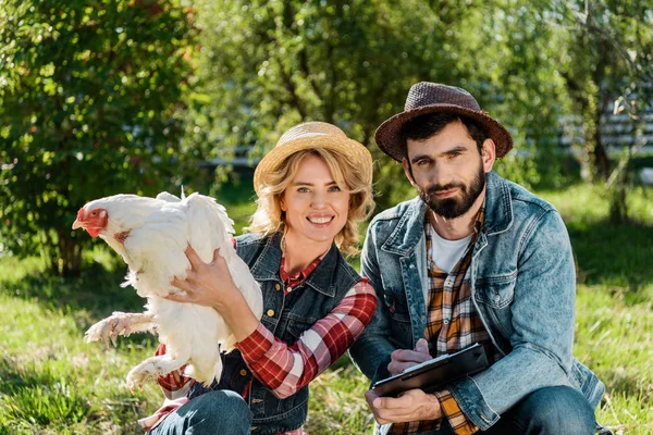 Mujer feliz sosteniendo pollo mientras su novio toma notas en portapapeles en la granja - foto de stock