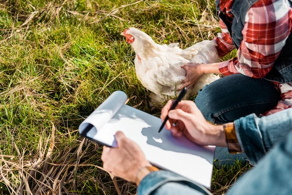 Vista parziale di agricoltore femminile che tiene pollo mentre il suo ragazzo prende appunti negli appunti all'aperto — Foto stock