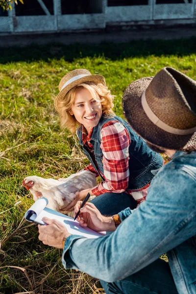 Веселая женщина держит курицу, пока ее парень делает заметки в бумагах на ферме — стоковое фото