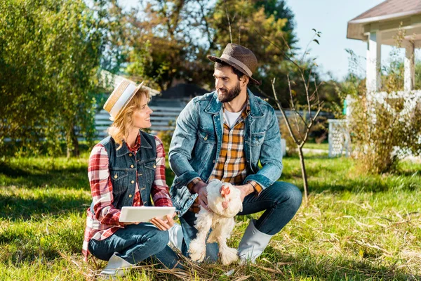 Lächelnder Landwirt hält Hühner, während seine Freundin auf dem Hof ein digitales Tablet benutzt — Stockfoto