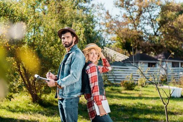 Erwachsenes Bauernpaar steht Rücken an Rücken mit digitalem Tablet und Klemmbrett auf der Ranch — Stockfoto