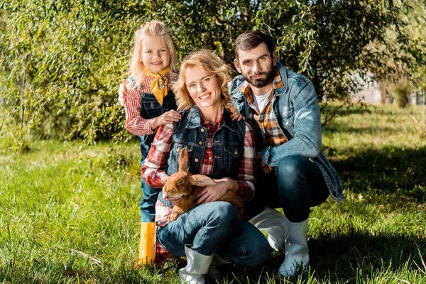 Cheerful farmer family with little daughter and brown rabbit sitting on grass outdoors — Stock Photo