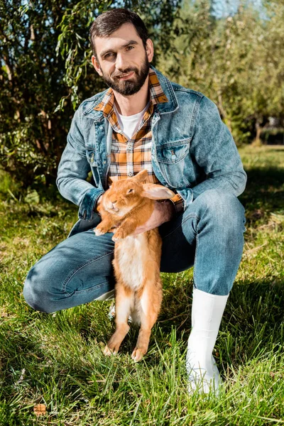 Heureux mâle agriculteur tenant lapin brun et regardant la caméra à l'extérieur — Photo de stock