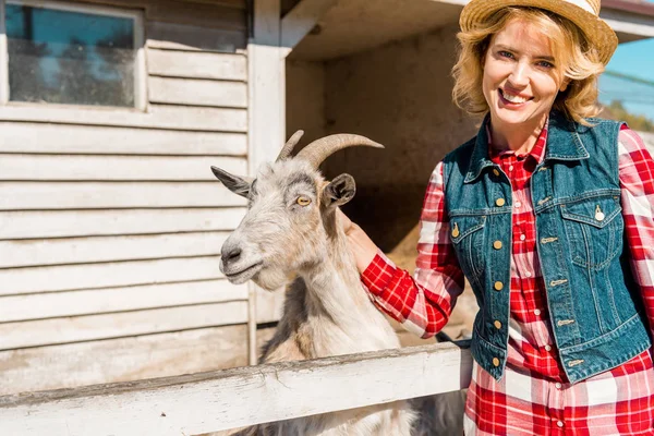 Mujer atractiva alegre tocando cabra cerca de valla de madera en la granja - foto de stock