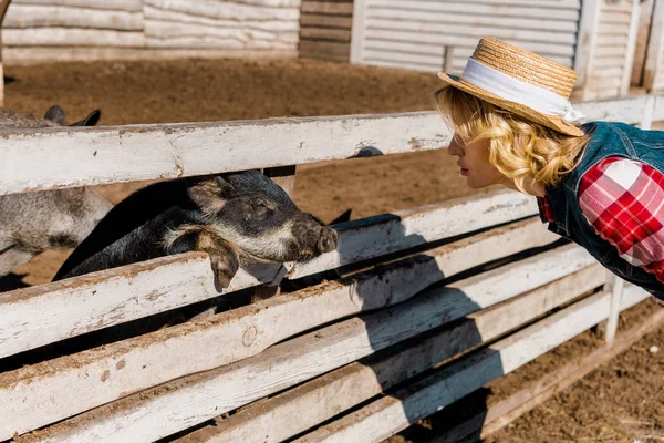 Side view of woman in straw hat looking at black piglet standing near wooden fence at farm — Stock Photo