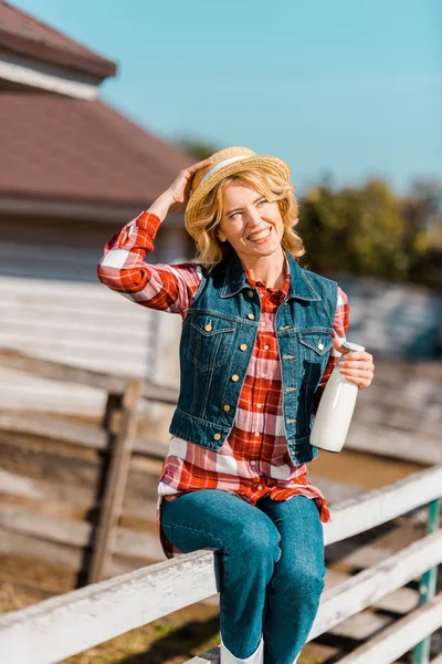 Happy female farmer holding bottle of milk and sitting on wooden fence at ranch — Stock Photo