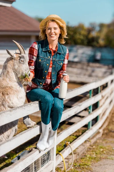 Smiling female farmer with milk bottle sitting on wooden fence and feeding goat at ranch — Stock Photo