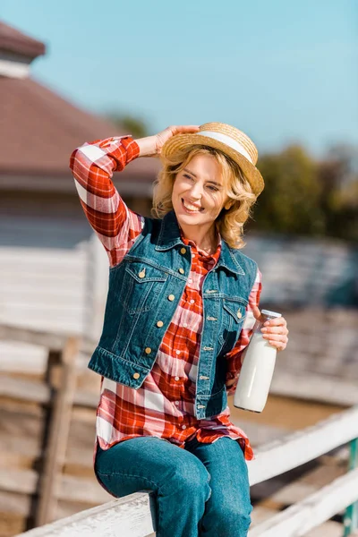 Enfoque selectivo de la sonriente agricultora sosteniendo botella de leche y sentada en una valla de madera en el rancho - foto de stock