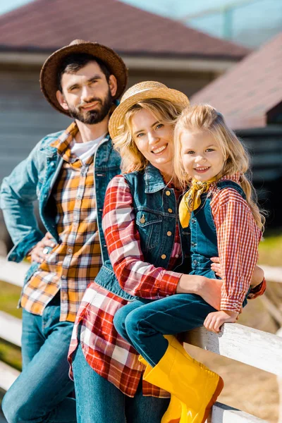 Selective focus of happy farmer family with little daughter sitting on wooden fence at ranch — Stock Photo