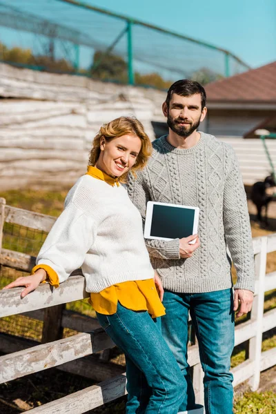 Sonriente pareja de agricultores mostrando tableta digital con pantalla en blanco cerca de valla de madera en la granja - foto de stock