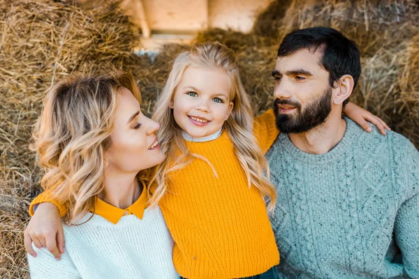 Pequeño niño abrazando a la madre y el padre mientras mira la cámara cerca de las pilas de heno en la granja - foto de stock
