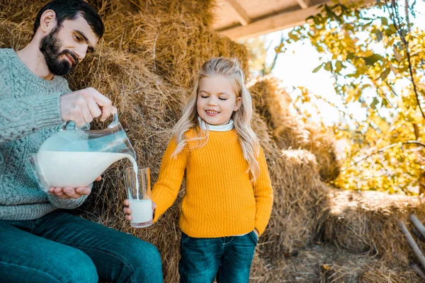 Adult female farmer pouring milk to little daughter near hay stacks at ranch — Stock Photo