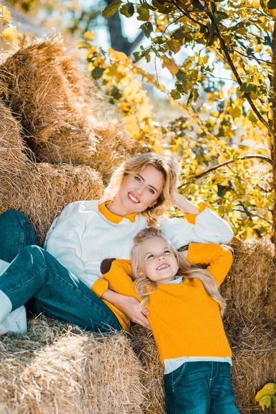 Sonriente mujer adulta posando con su hija pequeña en pilas de heno en la granja - foto de stock
