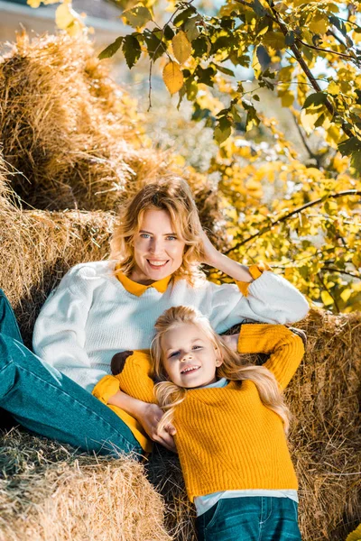 Mujer alegre posando con su hija pequeña en pilas de heno en la granja - foto de stock